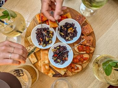 Wooden platter with three bowls of black rice salad, bruschetta, and crackers. Hands reach for food, white wine glasses surround the board.
