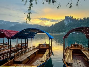 Drei bunte hölzerne Pletna-Boote am Bleder See bei Sonnenuntergang. Die Burg Bled thront auf einer bewaldeten Klippe im Hintergrund.