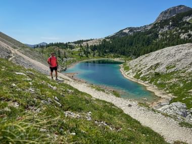 Wanderer in roter Jacke mit Blick auf türkisen Bergsee im Sieben-Seen-Tal, Julische Alpen, mit felsigen Hängen und bewaldeten Gipfeln.