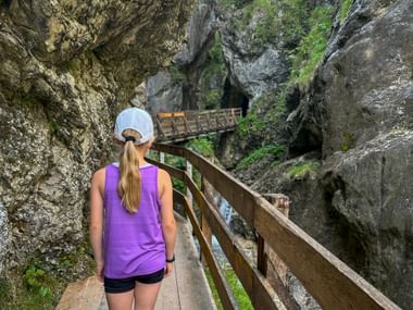Female hiker in purple top and white cap on wooden walkway through narrow rocky gorge with wooden bridges in Rosengartenschlucht near Imst.