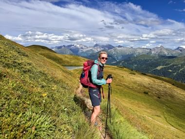 Wanderin mit Rucksack und Stöcken auf grasigem Berghang. Alpengipfel und kleiner See im Hintergrund unter blauem Himmel mit Wolken.