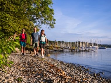 Three hikers with backpacks walking along a pebble shore at Starnberger See. A marina with sailboats and trees are visible in the background.