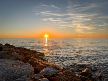 Sonnenuntergang über ruhigem Meer bei Cecina Mare in der Toskana. Sonne spiegelt sich im Wasser, felsige Küste im Vordergrund, orange-blauer Himmel.