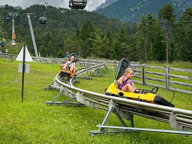 Two children riding colorful sleds on an alpine coaster track in Imst, with cable cars and forested mountains in the background.