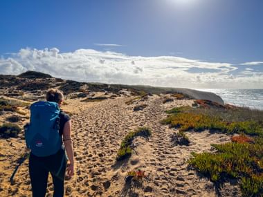 Hiker with blue backpack walking on sandy coastal path at Praia do Brejo Largo on the Rota Vicentina, with ocean views and dramatic clouds.