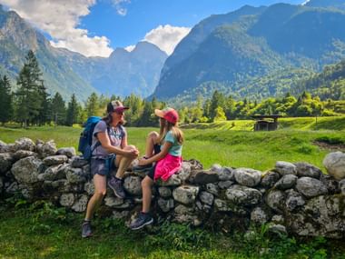 Zwei Wanderer sitzen auf einer Steinmauer in Trenta, Soča-Tal. Grüne Wiesen und bewaldete Berge der Julischen Alpen im Hintergrund.