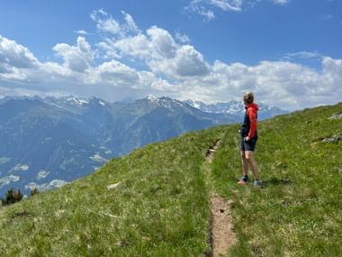 Wanderer in roter Jacke auf Bergpfad im Passeiertal bei Meran, mit grünen Wiesen und schneebedeckten Alpengipfeln unter blauem Himmel.