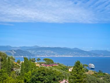 Panoramic view of the Italian Riviera coastline with villas, cypress trees, and a cruise ship on blue water. Mountains visible in the distance.