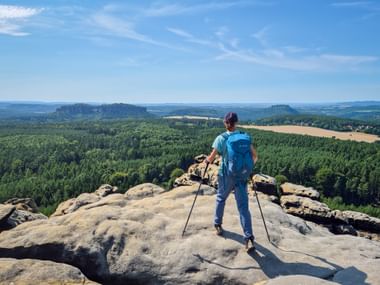 Hiker with blue backpack and trekking poles standing on rocky outcrop at Gohrisch viewpoint, overlooking forested landscape with fields.