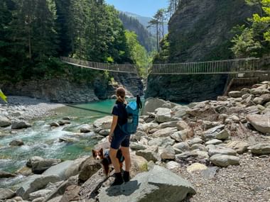 Female hiker with dog and blue backpack standing on rocks at Viamala Gorge, viewing suspension bridge over turquoise Hinterrhein river.