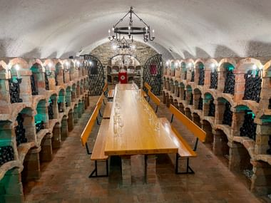 Vaulted wine cellar with brick arches, wooden tasting table, and wine bottles stored in alcoves. Iron chandelier hangs from ceiling.