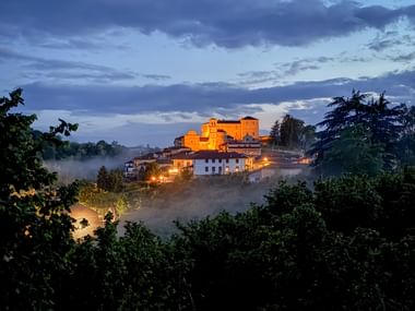 Illuminated hilltop village of Cravanzana in Piedmont at dusk, with a castle glowing orange above white houses, surrounded by fog and trees.