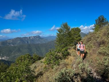 Two hikers walking on a mountain trail with panoramic views of forested valleys and mountains under a blue sky in Piedmont-Liguria region.