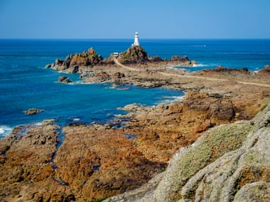 La Corbière Leuchtturm auf felsigem Vorsprung umgeben von blauem Meer. Zerklüftete Küstenfelsen im Vordergrund unter blauem Himmel.