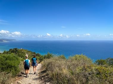 Zwei Wanderer mit Rucksäcken auf einem Erdweg am Monte Piccaro mit Blick auf das blaue Mittelmeer und Küstenvegetation.