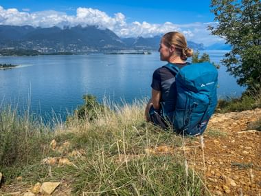 Wanderin mit blauem Rucksack sitzt am Hang der Rocca di Manerba und blickt auf den Gardasee mit Bergen und weißen Wolken im Hintergrund.