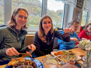 Four people enjoying lunch at a restaurant table with plates of food and drinks, large windows showing autumn landscape in background.