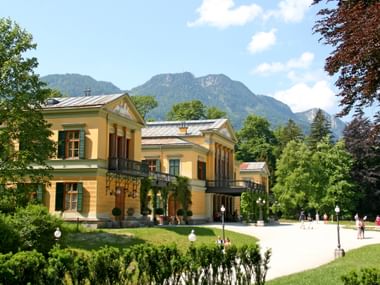 Yellow neoclassical Kaiservilla in Bad Ischl with columned entrance, surrounded by manicured gardens and visitors, against forested mountains.