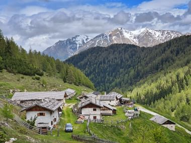 Traditionelles Almdorf Vallmingalm mit steinernen Dächern auf grünen Wiesen, umgeben von Wäldern und schneebedeckten Bergen.