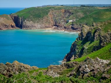 Grève de Lecq Bucht auf Jersey mit türkisem Wasser, Sandstrand, dramatischen Felsklippen und grünen Hügeln unter blauem Himmel.