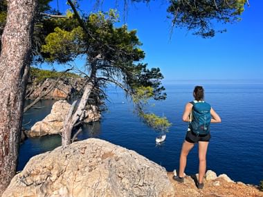 Hiker enjoys the view of a sailing boat and the rocky coast, surrounded by pine trees