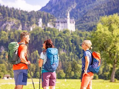 Three hikers in front of Neuschwanstein Castle, green nature, sunny weather, hikers smiling