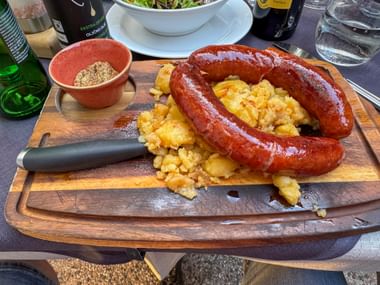 Krainer sausage arranged in a horseshoe shape on a wooden board with fried potatoes, accompanied by a small bowl of spices and a knife.