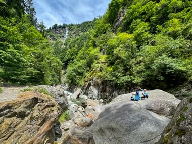 Two hikers resting on large rocks in Passer Gorge surrounded by lush green forest, with a waterfall visible in the background.