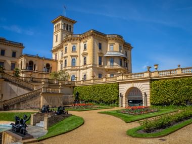 Osborne House, a grand Italianate palace with tower and curved facade, surrounded by formal gardens with manicured lawns and flower beds.