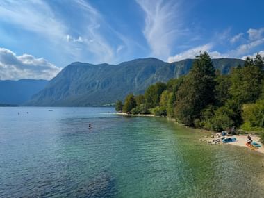 Klares türkisfarbenes Wasser des Bohinjer Sees mit bewaldeter Uferlinie und Julischen Alpen im Hintergrund. Kleiner Strand mit Badenden sichtbar.