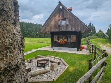 Dunkle Holzhütte mit steilem Dach in Pokljuka, mit Feuerstelle, grünen Wiesen, Holzzäunen und Wald im Hintergrund.