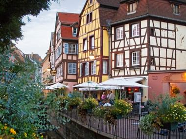 Bunte Fachwerkhäuser entlang eines Kanals im Viertel Klein-Venedig in Colmar. Eine Restaurantterrasse überblickt das Wasser, umgeben von Grün.