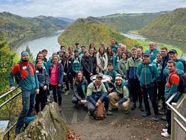 Large group of Eurohike team members in hiking gear posing at a scenic viewpoint overlooking the Danube River with forested hills and mountains.