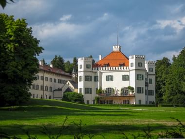 White Possenhofen Castle with red roof and green shutters, surrounded by manicured lawn and trees under a cloudy sky.