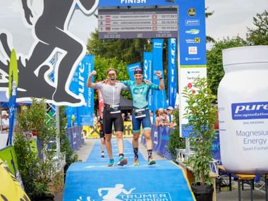 Two male athletes celebrating on podium at Trumer Triathlon 2026 finish line, wearing race gear and raising arms in victory under blue arch.