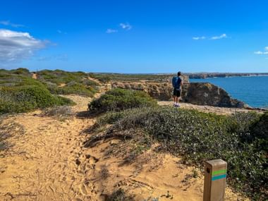 Wanderer mit Rucksack auf sandigem Küstenweg mit Holzwegweiser, Blick auf Klippen und blaues Meer an der Rota Vicentina, Portugal.