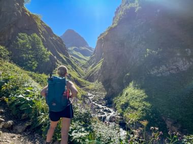 Wanderin mit Rucksack steht an einem Bergbach im Sperrbachtobel, umgeben von steilen Felswänden und grüner Vegetation unter blauem Himmel.