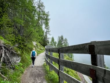 Hiker with blue backpack walking on gravel mountain trail with wooden fence railing, surrounded by green forest and misty valley views.