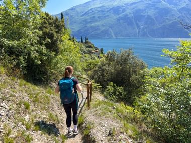 Wanderin mit türkisfarbenem Rucksack auf Bergweg mit Blick auf den Gardasee, umgeben von mediterraner Vegetation und Zypressen.