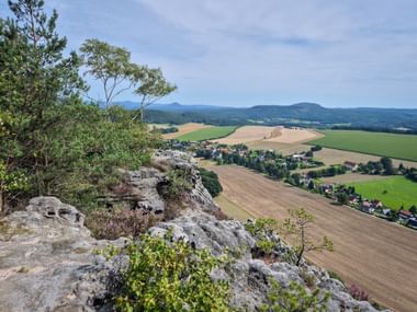 View from Papststein rock formation showing patchwork fields, a village, and distant mountains under blue sky along the Malerweg trail.