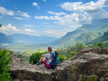 Wanderin mit blauem Rucksack sitzt auf Felsvorsprung mit Blick auf grünes Tal in Südtirol mit Bergen und blauem Himmel.