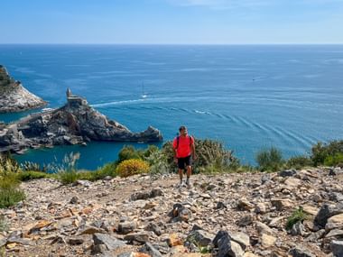 Hiker in red jacket on rocky trail overlooking Porto Venere bay with church on promontory, blue Mediterranean Sea, and sailboats.