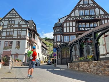 Wanderer mit Rucksack auf Kopfsteinpflasterstraße, umgeben von traditionellen deutschen Fachwerkhäusern unter blauem Himmel.