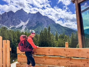 Wanderin in roter Jacke mit Rucksack auf Holzterrasse in Mieders, Koppeneck. Alpengipfel und Wald unter blauem Himmel mit Wolken sichtbar.