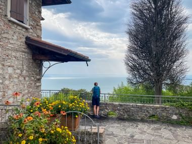 Person in blauer Jacke steht an Steinterrasse mit Blick auf den Gardasee. Bunte Blumen im Vordergrund, Steingebäude und Baum sichtbar.