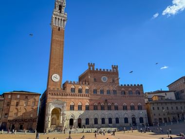Piazza del Campo in Siena mit dem mittelalterlichen Palazzo Pubblico und seinem hohen Torre del Mangia unter blauem Himmel. Menschen versammeln sich auf dem Platz.
