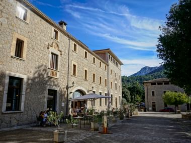 Stone courtyard of Monastery Lluc with outdoor café, historic buildings, and Tramuntana Mountains in the background under blue sky.