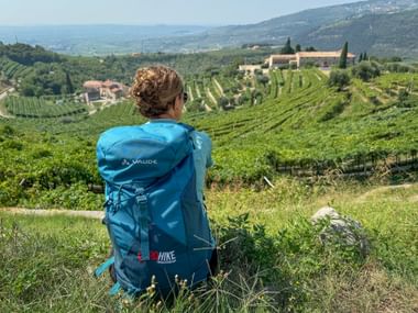 Hiker with blue backpack sitting in grass overlooking terraced vineyards and hills near Verona and Lake Garda, with cypress trees and buildings.