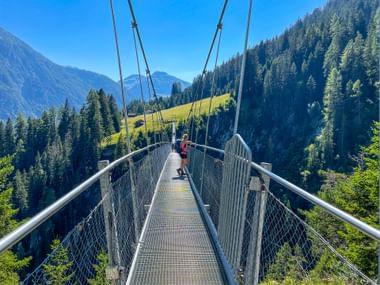 Wanderer überquert eine Hängebrücke in Holzgau mit Berggipfeln und bewaldeten Hängen in den Allgäuer und Lechtaler Alpen unter blauem Himmel.