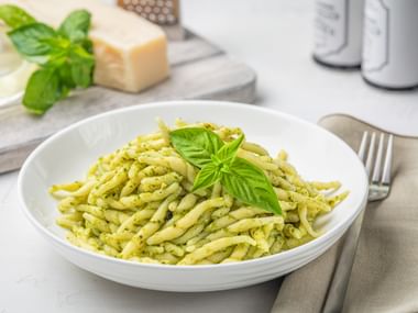 White bowl of trofie pasta coated in green pesto Genovese, garnished with fresh basil. Parmesan cheese and grater visible in background.